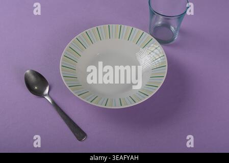 Minimalistisches Stillleben mit leerer weißer Schüssel, metallischem Löffel und Glas vor massivem violettem Hintergrund. Shot verfügt über eine pastellfarbene Farbpalette und ist weich Stockfoto