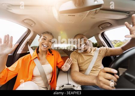 Glückliches junges afroamerikanisches Paar, das im Auto winkt und während der Reise begrüßt Stockfoto