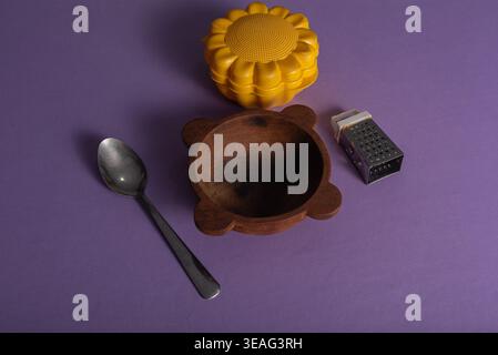 Stillleben zeigt Lebensmittel, die vor violettem Hintergrund isoliert sind. Holzschale, Metalllöffel, Reibe, strukturierter Behälter in Sonnenblumenform. Matt Stockfoto