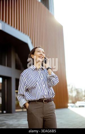 Eine fröhliche Koreanerin steht vor einem modernen Gebäude und unterhält sich mit einem hellen Lächeln am Telefon. Die Sonne scheint warm oben. Stockfoto