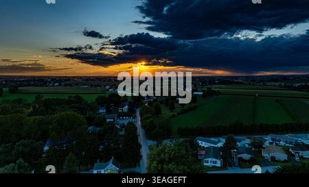 Die Sonne geht hinter einer Wolkenlinie unter und wirft orange und gelbe Farben über den Himmel. Darunter erstrecken sich Felder und eine kleine Stadt mit Häusern ist zu sehen. Bäume säumen die Straßen. Stockfoto