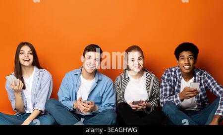Gruppe von vier Freunden, die Freizeit genießen, während sie Smartphones gegen eine orangefarbene Wand verwenden Stockfoto