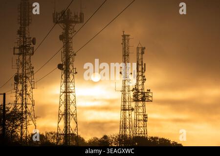 Telekommunikationstürme und Mobiltelefonmasten, die sich vor einem strahlenden Sonnenuntergangshimmel mit sichtbaren Antennen und Sendeanlagen darstellen Stockfoto