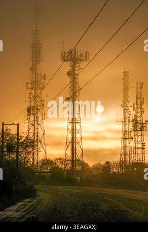 Telekommunikationstürme und Mobiltelefonmasten, die sich vor einem strahlenden Sonnenuntergangshimmel mit sichtbaren Antennen und Sendeanlagen darstellen Stockfoto