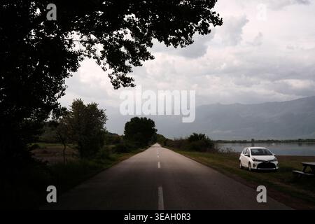 Ein einsames weißes Auto parkt auf der grasbewachsenen Schulter einer langen, leeren Asphaltstraße, die in Richtung trübe Berge neben einem ruhigen See führt Stockfoto
