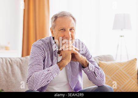 Der lächelnde Seniorenmann sitzt tagsüber in einem hellen Wohnzimmer mit den Händen auf der Couch Stockfoto