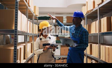 Mann und Frau organisieren Produkte in Regalen und Regalen, schauen sich die Warenbestände auf Laptops und Tablets an. Team von Arbeitern mit Overalls, die Waren im Lagerraum arrangieren, Versand bestellen. Stockfoto