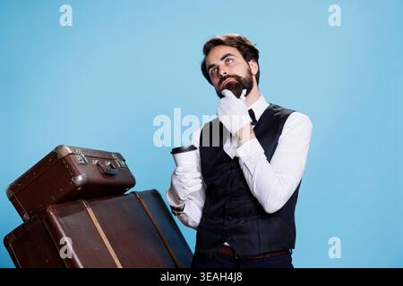 Ein aufmerksamer Portier trinkt Kaffee im Studio und steht neben dem Gepäck mit Anzug und Krawatte. Junger Mann mit Pagen-Job, der eine Tasse kaltes Bier genießt und selbstbewusst posiert. Stockfoto