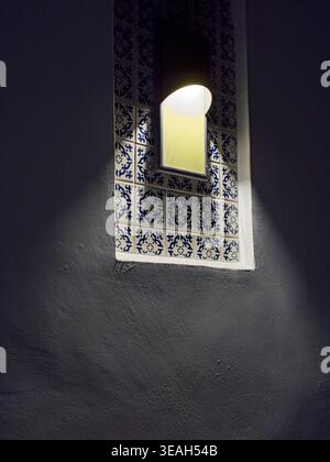 Beleuchtetes gemauertes Fenster mit blau-weißen Porzellanfliesen auf grauer Stuckwand Stockfoto
