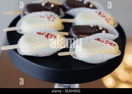 Weiße Schokolade Pops Auf Schwarzem Stand, Elegante Hochzeit Dessert Display Durch Konditor, Weiße Schokolade Beschichtung Mit Roten Sprinkle Garnish, Papierbecher Stockfoto