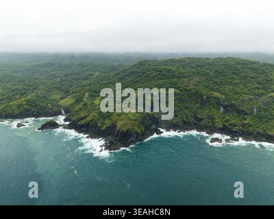 Blick aus der Vogelperspektive auf Bukit Pengilon mit üppigen grünen Hügeln, felsigen Klippen und einem kleinen Wasserfall, der in das türkisfarbene Meer in Kecamatan Tepus, D, stürzt Stockfoto