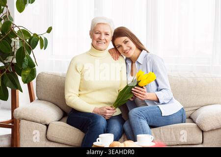 Zwei Frauen teilen sich einen freudigen Moment mit gelben Blumen auf einem gemütlichen Sofa in einem hellen Zimmer Stockfoto