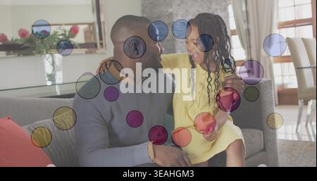 Lachender Vater, der ein Mädchen auf grauem Sofa zu Hause in hellgrauem Pullover-gelbem Kleid mit Überzügen hält Stockfoto