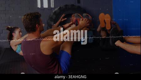 Der darstellende Mann trägt ein kastanienbraunes Tanktop und blaue Shorts, der V-Sit auf dem Fitnessboden hält, Traktorreifen Stockfoto