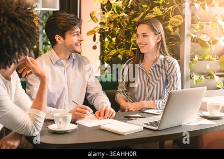 Teammitglieder arbeiten während einer Arbeitssitzung in einem gemütlichen Café mit Pflanzen und einem Laptop zusammen Stockfoto