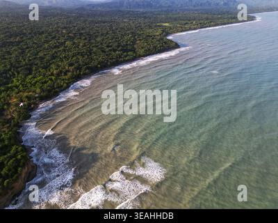 Blick aus der Vogelperspektive auf die üppige tropische Küste, wo der dichte Dschungel auf das türkisfarbene Karibische Meer mit den Wellen und den fernen Bergen Palomino, La, trifft Stockfoto
