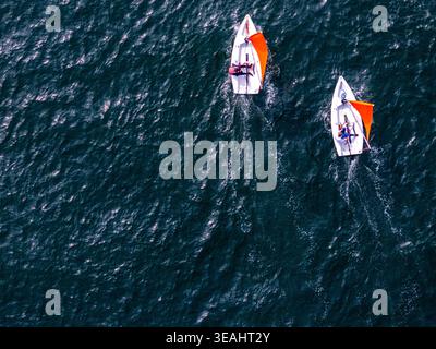 Aus der Vogelperspektive von zwei kleinen weißen Segelbooten mit orangefarbenen Segeln und Personen an Bord, die über dunkelblaues, plätscherndes Wasser mit weißen Wachen segeln. Stockfoto
