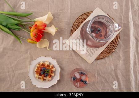 Ein Glas Eistee steht auf einem Korbgeflecht neben einem Teller mit Waffeln, umgeben von einem Tulpenstrauß und einem Buch. Stockfoto