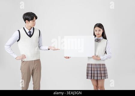 Eine junge asiatische Frau in Schuluniform hält ein leeres Schild, während ein junger asiatischer Mann in Schuluniform ihr eine Geste gibt. Stockfoto