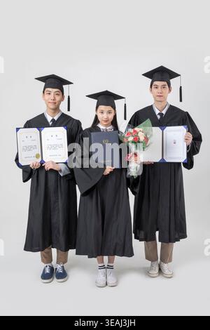 Drei Absolventen unterschiedlichen Alters stehen zusammen, halten ihre Diplome und lächeln stolz in ihren Kappen und Gewändern. Stockfoto