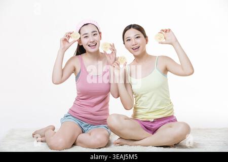 Zwei junge Frauen sitzen auf dem Boden, jede hält ein Stück Zitrone vor ihren Gesichtern, lächelt und blickt in die Kamera. Stockfoto