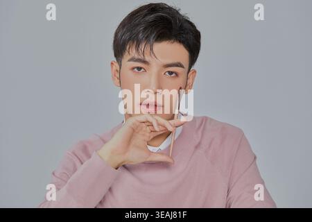 Ein junger asiatischer Mann in einem rosafarbenen Pullover hält ein Smartphone an seinem Ohr und blickt mit besorgtem Ausdruck nach oben. Stockfoto
