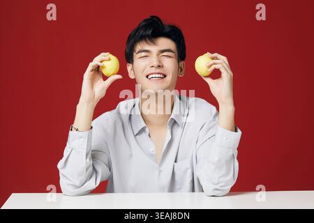 Ein junger Mann sitzt an einem weißen Tisch, hält zwei Zitronen und lächelt in die Kamera. Stockfoto