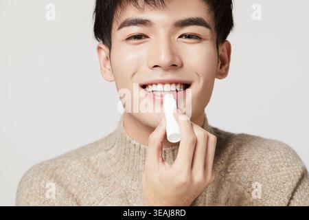Ein junger Mann in einem beigefarbenen Pullover trägt Lippenbalsam auf seine Lippen. Stockfoto