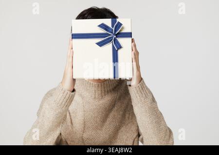 Eine junge Frau hält eine Geschenkverpackung vor ihrem Gesicht, ihre Augen hinter dem Geschenkpapier versteckt. Stockfoto