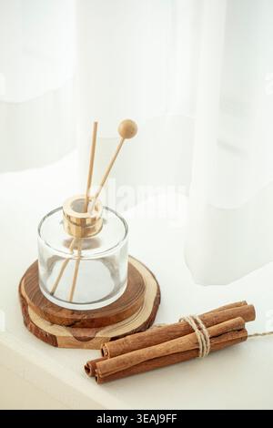Ein Glasrohr-Diffusor sitzt auf einem hölzernen Untersetzer, umgeben von einem Bündel Zimtstangen auf einem weißen Tisch. Stockfoto