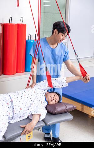 Ein junger asiatischer Physiotherapeut, der einer älteren Patientin bei einer Rehabilitationsübung auf einem Krankenhausbett hilft. Stockfoto
