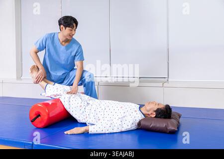 Ein männlicher Patient liegt auf einer bettähnlichen Matte mit einem medizinischen Fachpersonal in blauer Uniform, das ihm bei Dehnungsübungen hilft. Stockfoto