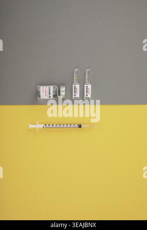 Eine Spritze und mehrere Durchstechflaschen mit COVID-19-Impfstoff sind auf einem gelben und grauen Hintergrund angeordnet. Stockfoto