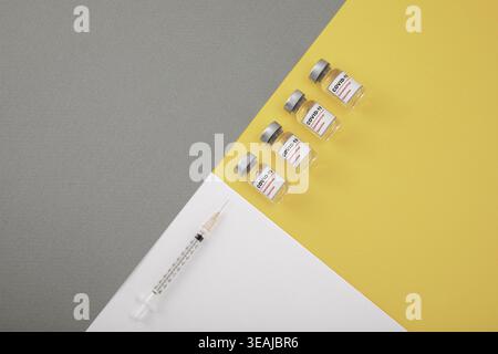 Vier COVID-19-Impfstoffflaschen und eine Spritze liegen auf einer grauen und gelben Oberfläche. Stockfoto