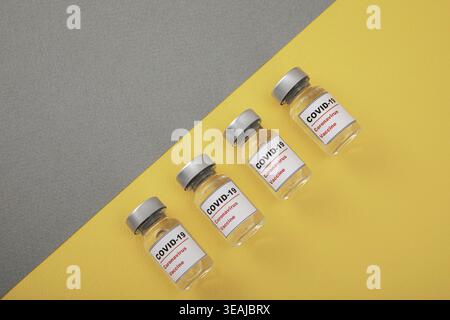 Vier leere COVID-19-Impfstofffläschchen sind auf einer gelben und grauen Oberfläche angeordnet. Stockfoto