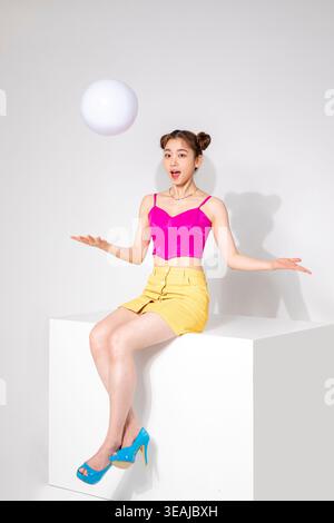 Eine junge Frau in rosa Oberteil und gelbem Rock sitzt auf einem weißen Würfel und fängt einen weißen Ball in der Luft. Stockfoto