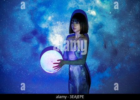 Eine junge Frau steht an einem Mondstrand und hält einen glühenden Mond in ihren ausgestreckten Händen. Stockfoto