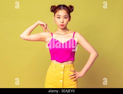 Eine junge asiatische Frau mit Brötchen und farbenfroher Kleidung steht selbstbewusst in einem Studio-Setting und posiert mit einer Hand auf der Hüfte. Stockfoto
