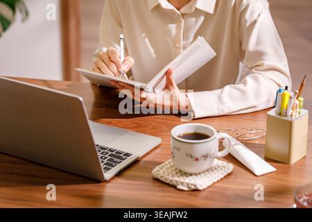Eine Frau sitzt an einem hölzernen Schreibtisch und schreibt in einem Notizbuch, während sie in einem ruhigen, natürlich beleuchteten Büro an einem Laptop arbeitet. Stockfoto