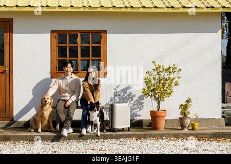 Ein junges Paar sitzt an einem sonnigen Morgen auf einer Bank vor ihrem Haus, umgeben von ihren Hunden. Stockfoto