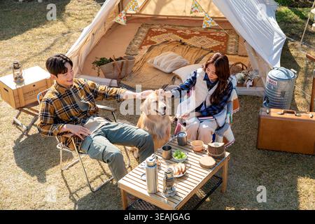 Ein Mann und eine Frau sitzen auf einem Campingplatz und streicheln einen goldenen Retriever, während sie draußen essen. Stockfoto
