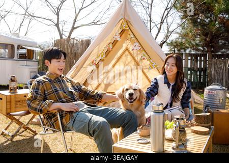 Ein Mann und eine Frau sitzen am Lagerfeuer, lachen und streicheln einen goldenen Hund auf einem sonnigen Campingplatz. Stockfoto