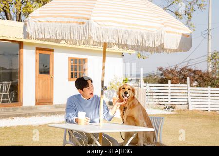 Ein junger Mann und ein goldener Retriee-Hund sitzen zusammen unter einem gestreiften Regenschirm in einem sonnigen Garten. Stockfoto