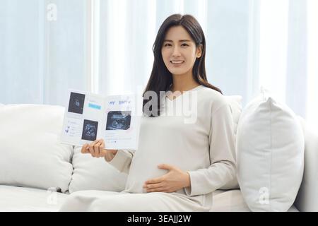 Eine junge Frau sitzt auf einer Couch und hält ein Babybuch mit Sonogrammen und Notizen. Stockfoto