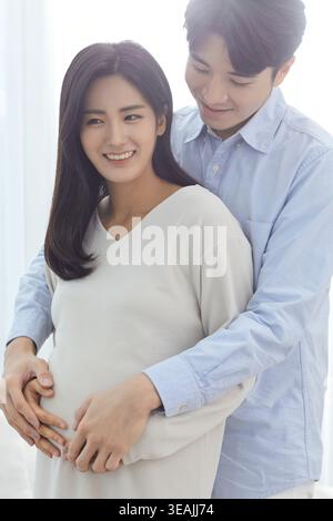 Eine junge asiatische Frau in einem weißen Pullover wird von ihrem Partner gewiegt, der ihren schwangeren Bauch hält. Stockfoto