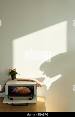 Ein warmes, goldenes Licht beleuchtet ein frisch gebackenes Brot in einem kleinen, weißen Elektroofen, der auf einem Holztisch in einer sonnigen Küche steht. Stockfoto