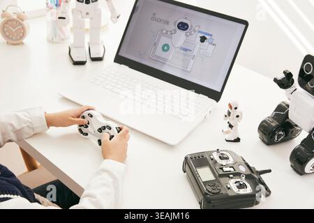 Eine Person sitzt an einem Schreibtisch und steuert einen kleinen Roboter mit einer Fernbedienung, während ein Laptop mit KI-Schulungsbildschirm vor ihnen geöffnet ist. Stockfoto