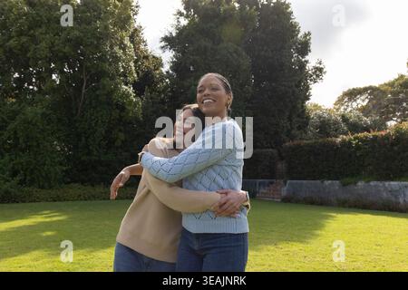 Verschiedene weibliche Partner, die sich auf gepflegtem Hinterhofrasen einlassen, Pullover und Armbanduhr tragen Stockfoto