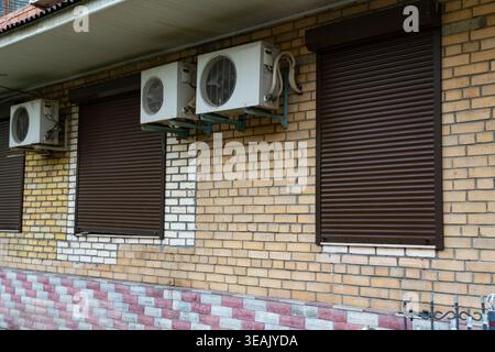 Mehrere Klimaanlagen, die an einer Ziegelwand mit geschlossenen Fensterläden montiert sind. Städtisches Infrastrukturkonzept in Bezug auf Kühlsysteme und bui Stockfoto