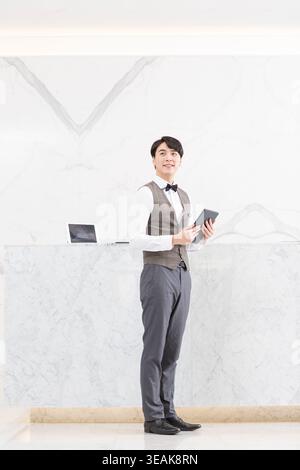 Ein junger männlicher Rezeptionist mit einem Tablet, der an einem modernen Marmorrezeptionstresen in einer eleganten Bürolobby steht. Stockfoto